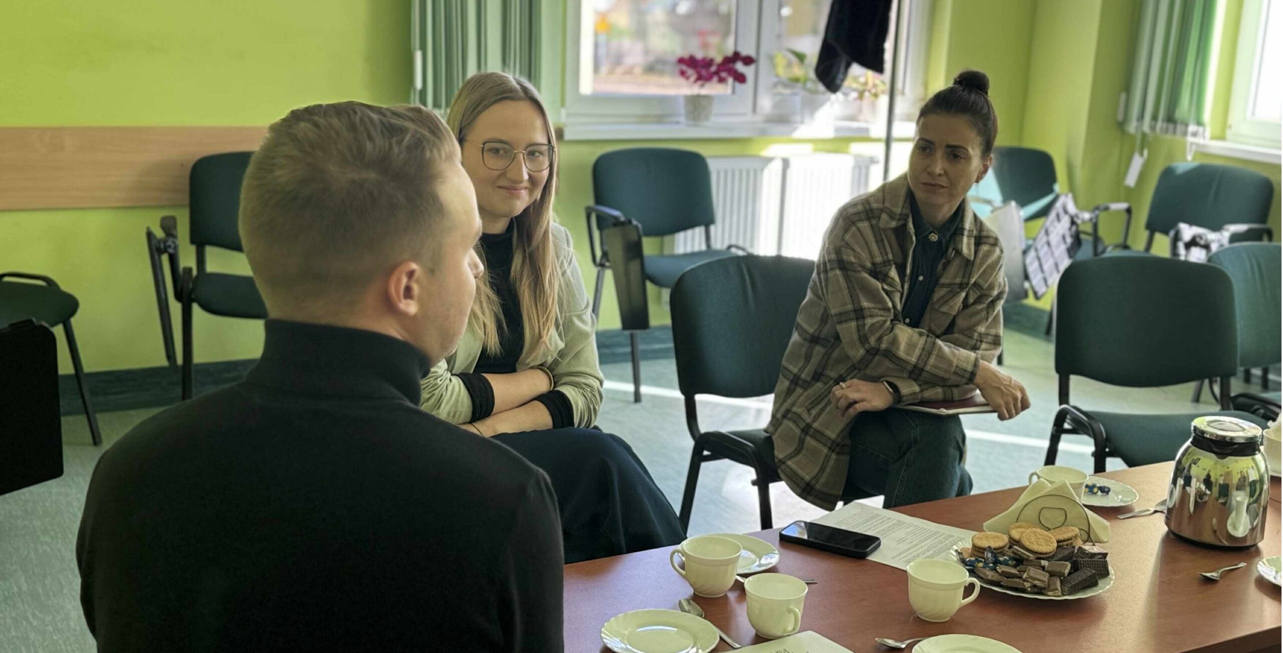 2 kobiety i mężczyzna siedzą przy stole, na którym stoją filiżanki i talerze. Rozmawiają.